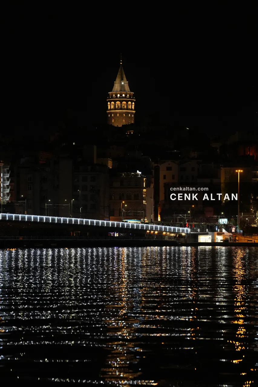 Gece Galata Kulesi ve önündeki aydınlatılmış köprü, suda ışık çizgileri