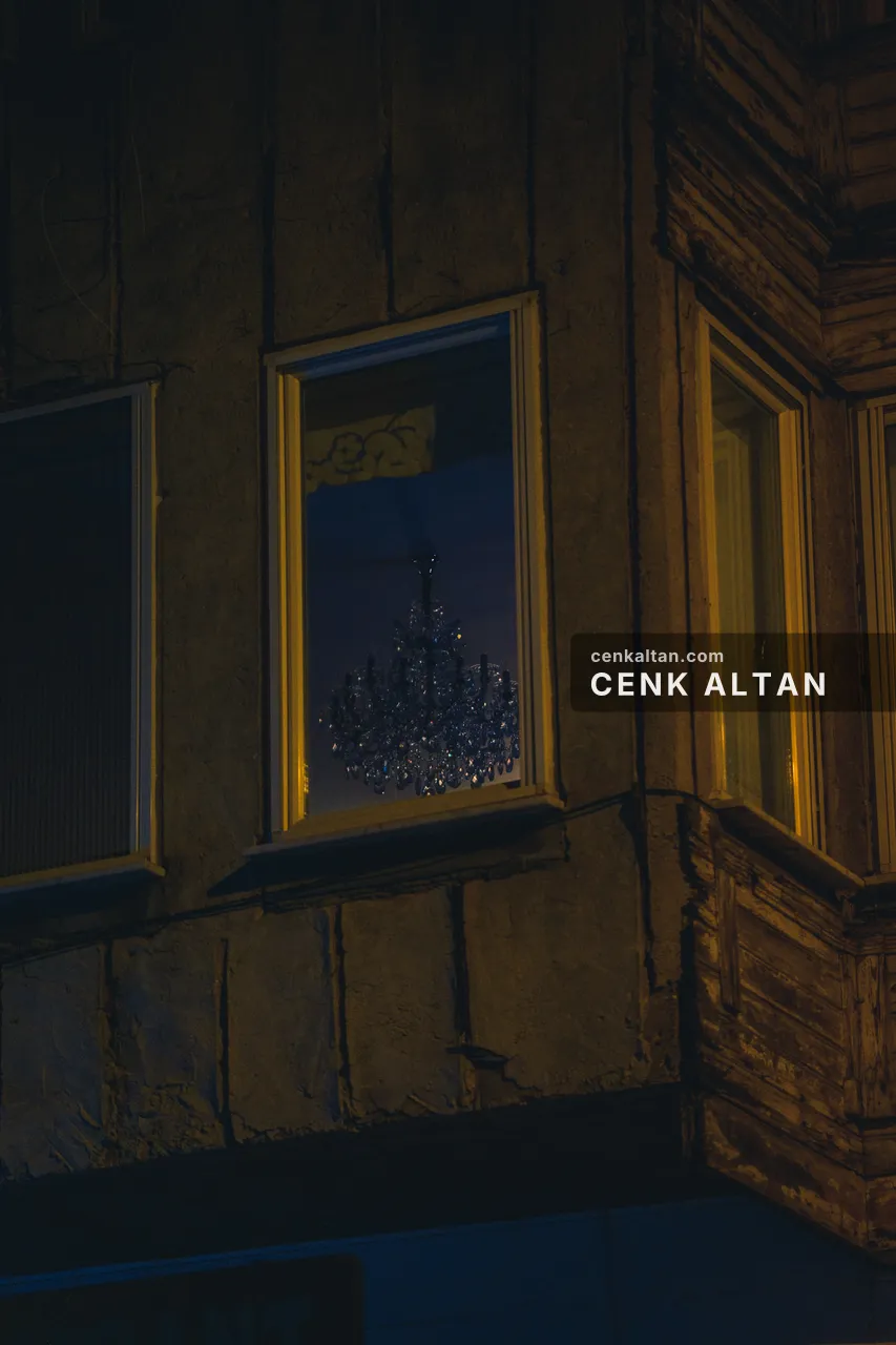 Gece, eski bir binanın sarı çerçeveli penceresinde kristal avize; cephede soyulmuş sıva ve ahşap cumba