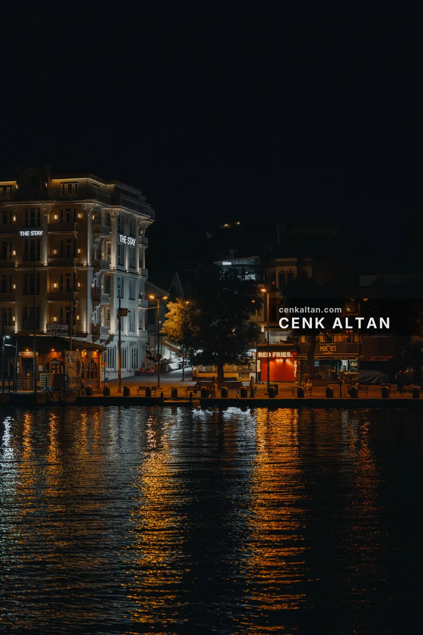 Gece, deniz üstünden Ortaköy rıhtımı: aydınlatılmış beyaz otel binası, ağaçlar, kapalı büfeler ve suda sarı yansımalar