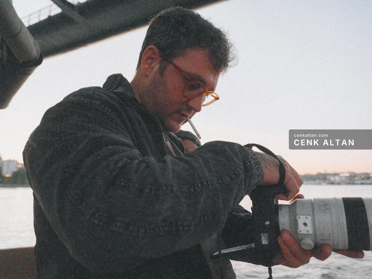 Sabah ışığında beyaz teleobjektifli fotoğraf makinesini kontrol eden gözlüklü adam; ağzında sigara, arkada köprü kirişi ve su