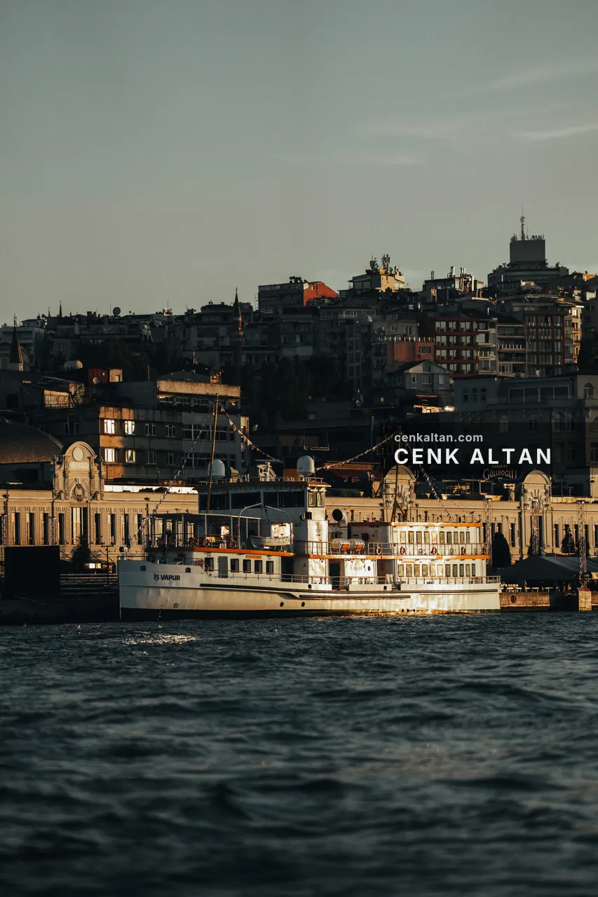 Sabah ışığında rıhtıma bağlı beyaz, eski tip bir vapur; arkada kemerli taş binalar ve tepeye tırmanan mahalle