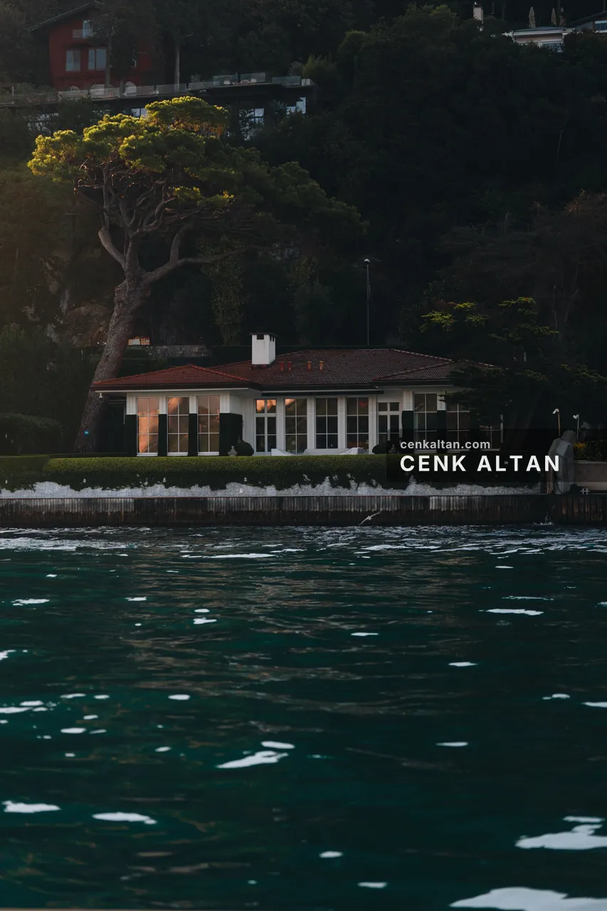 Sık ağaçlı yamacın eteğinde geniş pencereli, kiremit çatılı beyaz kıyı evi; solda fıstık çamı, önde köpüklü yeşil su