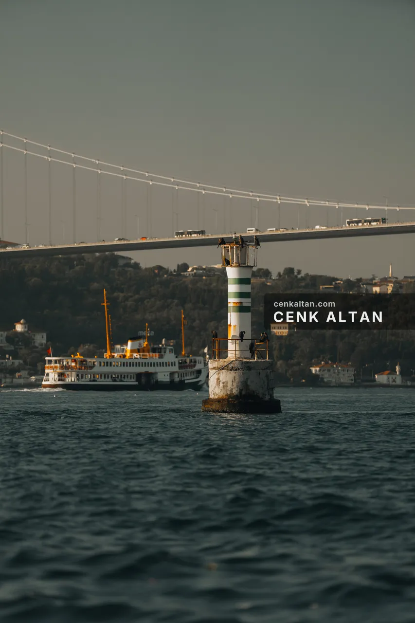 Boğaziçi Köprüsü'nün altında çizgili fener kulesi ve yanından geçen şehir hatları vapuru