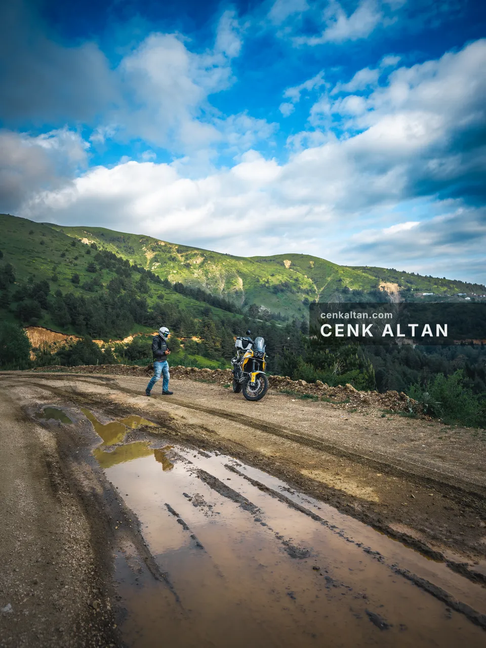 Yeşil tepeler ve bulutlu gökyüzü altında çamurlu yolda motosikletli