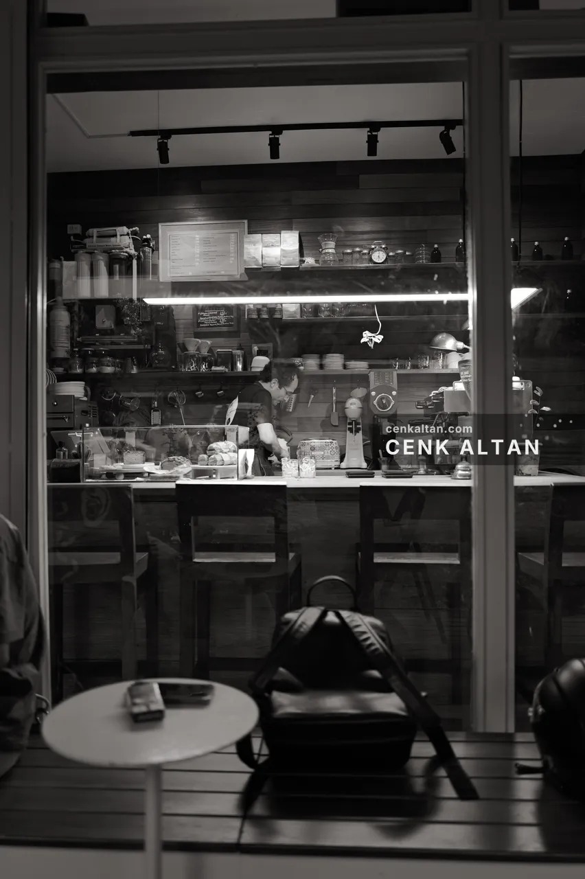 Siyah beyaz: cam arkasında çalışan barista, önde boş masa ve çanta