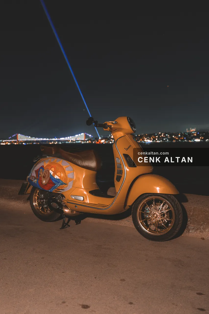 Gece deniz kenarında sarı Vespa, karşıda şehir ışıkları
