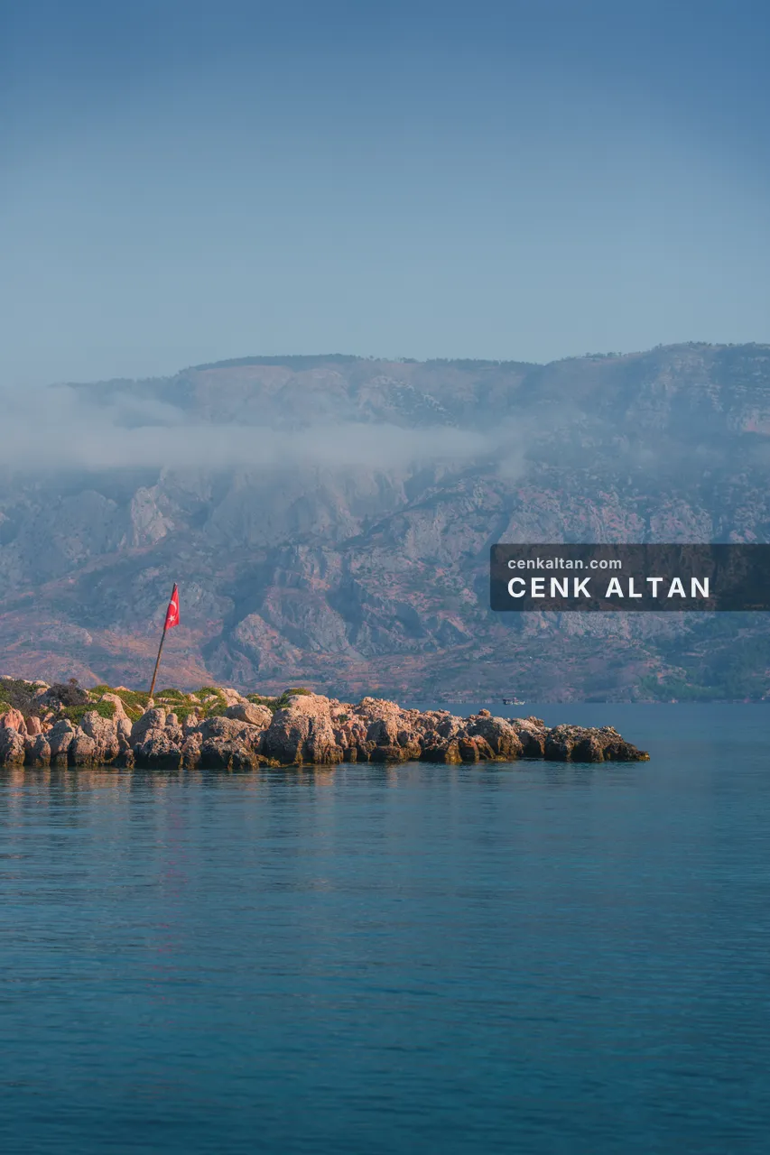 Denizde küçük bir kaya adacığı ve üstünde bayrak, arkada sisli dağlar
